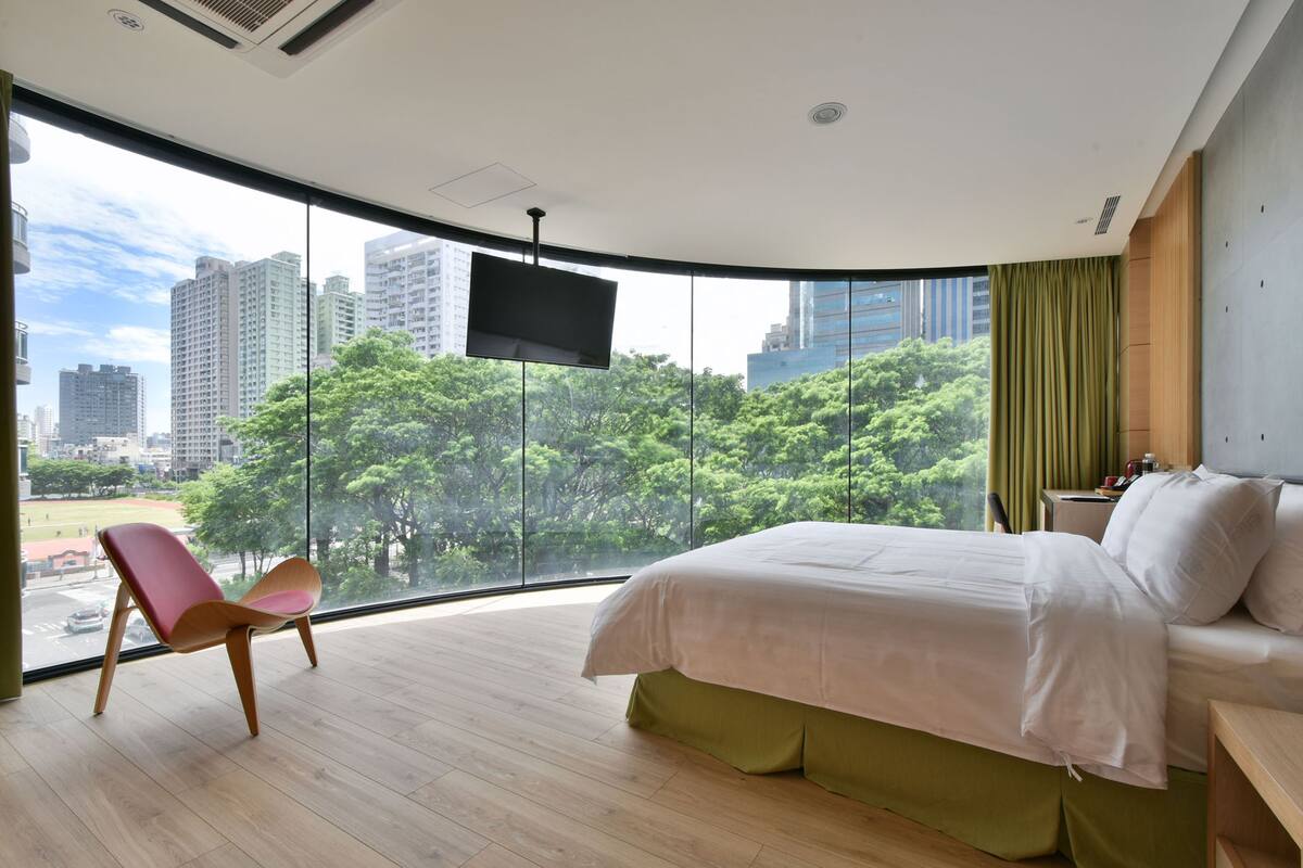 A modern hotel room with a large window, a bed, a chair, and a flat-screen TV.