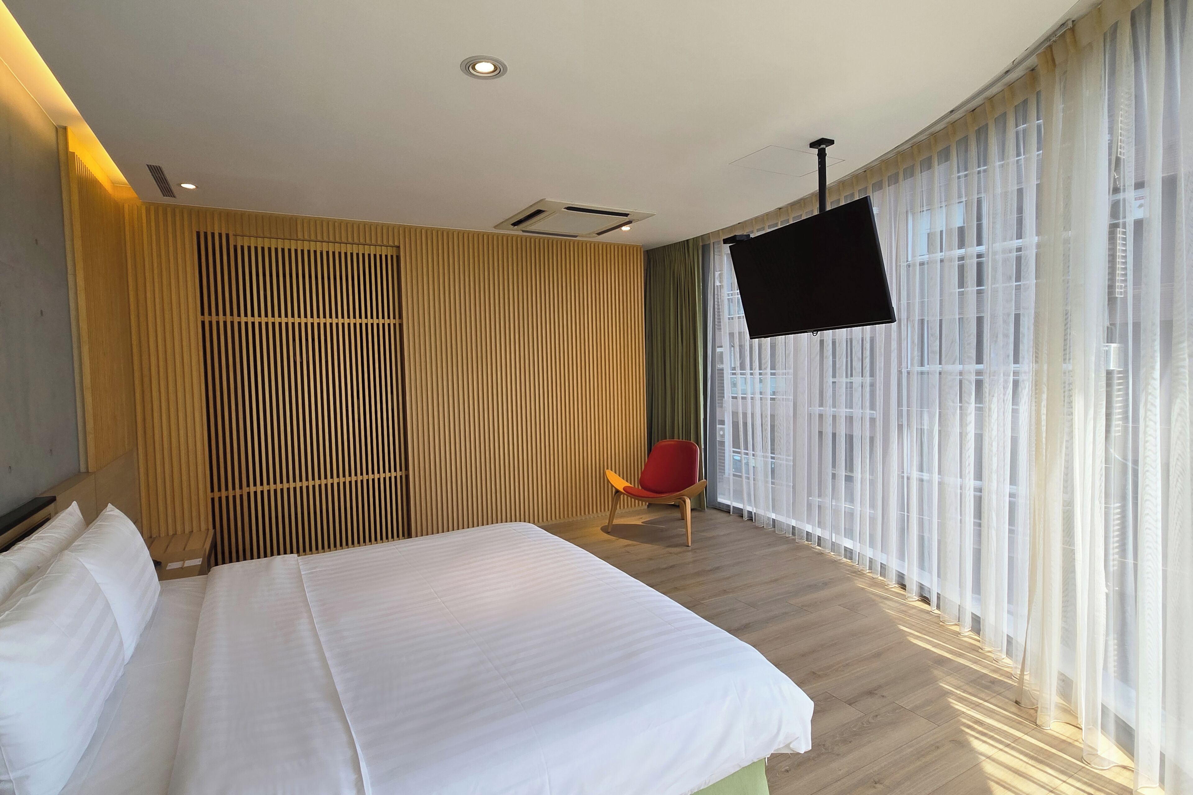 Ein Hotelzimmer mit einem großen Bett, einem an der Wand befestigten Fernseher und einem Fenster mit transparenten Vorhängen.