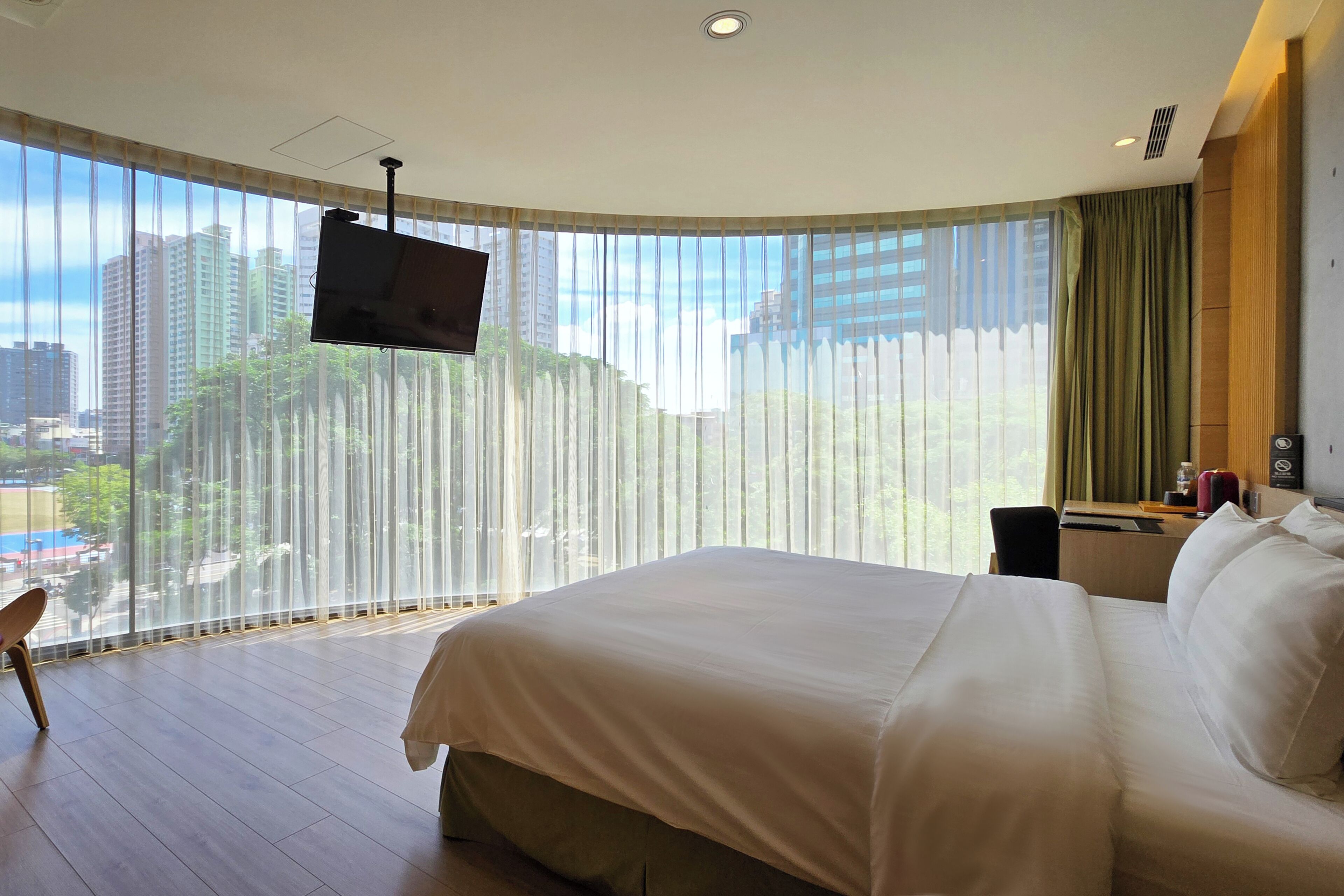 Ein Hotelzimmer mit einem großen Bett, einem an der Wand montierten Fernseher, einem Schreibtisch mit Stuhl und Blick auf die Stadt durch leichte Vorhänge.