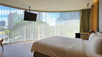 Ein Hotelzimmer mit einem großen Bett, einem an der Wand montierten Fernseher, einem Schreibtisch mit Stuhl und Blick auf die Stadt durch leichte Vorhänge.