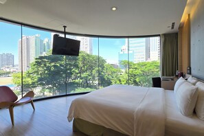 A hotel room with a large bed, a TV mounted on the wall, and a view of a cityscape through large windows.