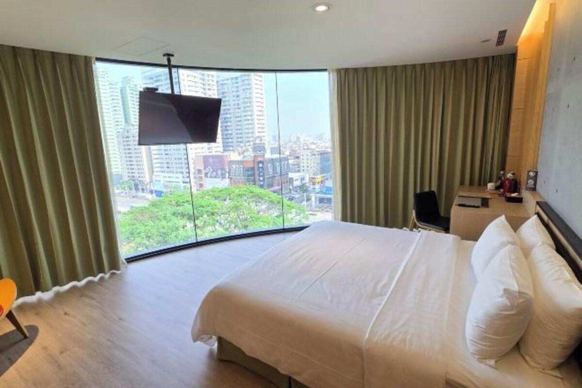 A hotel room with a large bed, a TV mounted on the wall, and a view of the city through large windows.