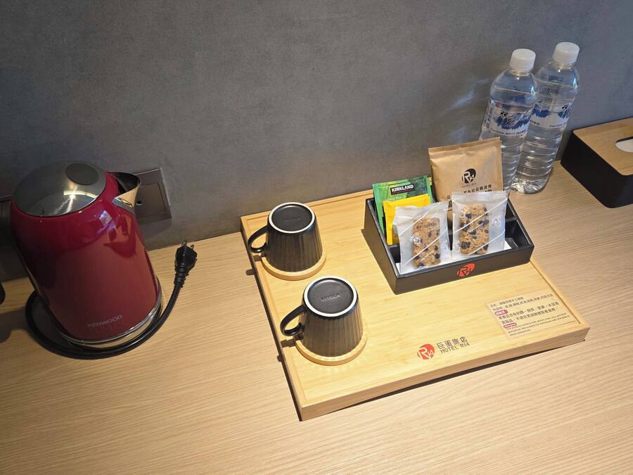 A hotel room with a red kettle, two cups, a tray with snacks and water bottles.