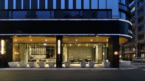 Un hotel modern amb façana corba, zona d'estar àmplia amb jardineres i un restaurant visible a l'interior.