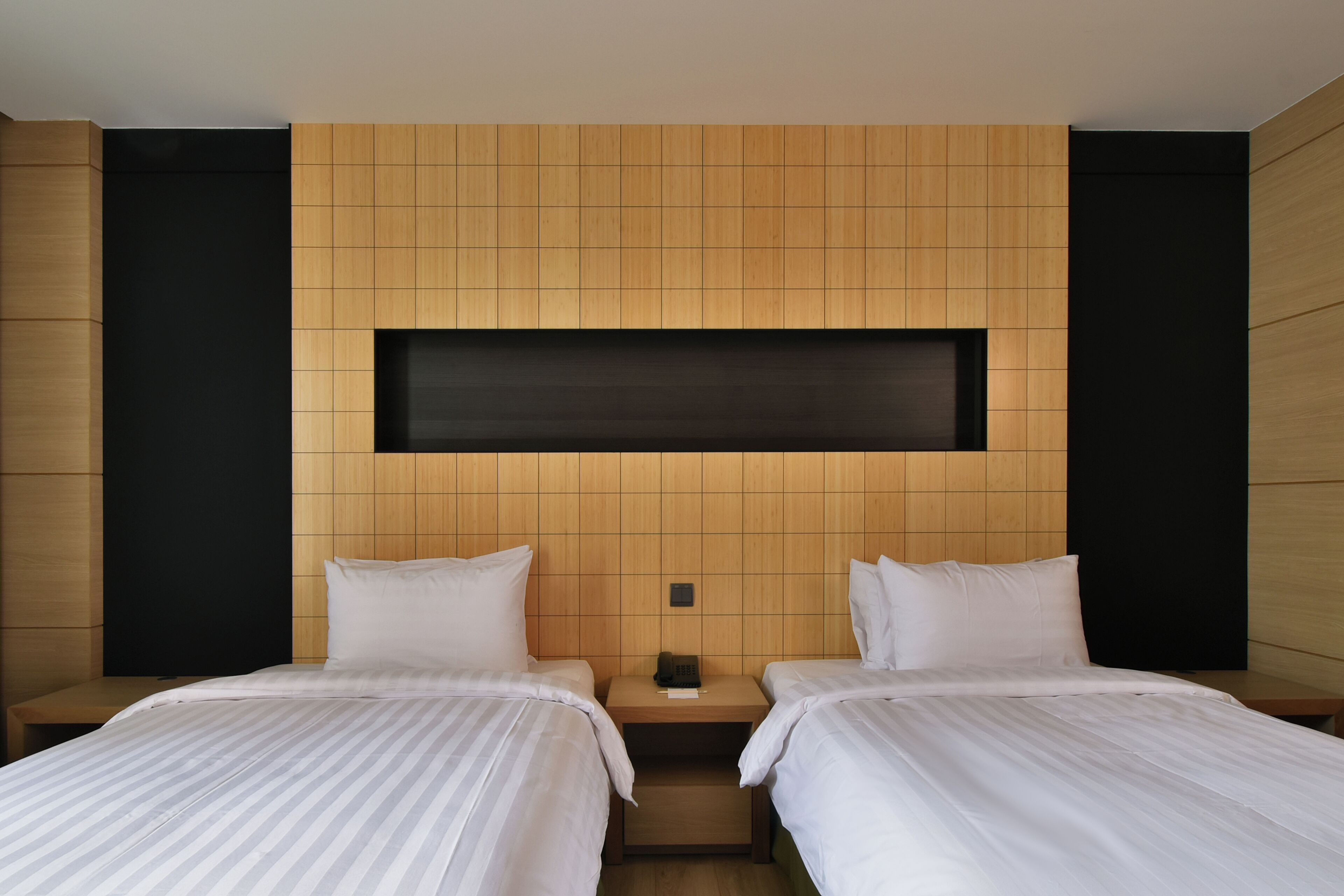 A hotel room with two beds, a wooden headboard, a small bedside table, and a black shelf above the beds.