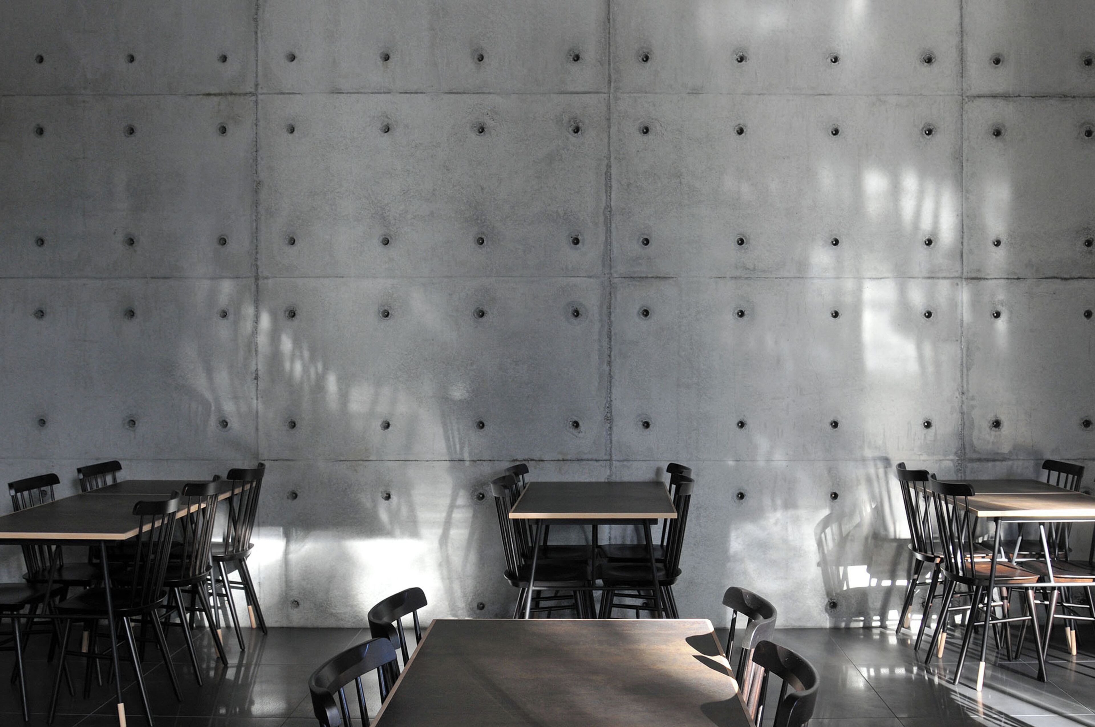 Un comedor con mesas y sillas, una pared de concreto con hendiduras circulares, y luz natural proyectando sombras.