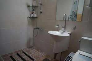 Un baño con lavamanos blanco, espejo, ducha e inodoro. Hay repisas con artículos de aseo y un panel de control en el lavamanos.