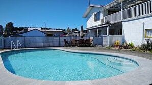 Ein Pool mit gewölbter Kante, umgeben von einer gepflasterten Terrasse. Liegestühle und ein Tisch sind für das Essen im Freien bereitgestellt. Ein weißes Gebäude mit Balkon grenzt an den Pool.