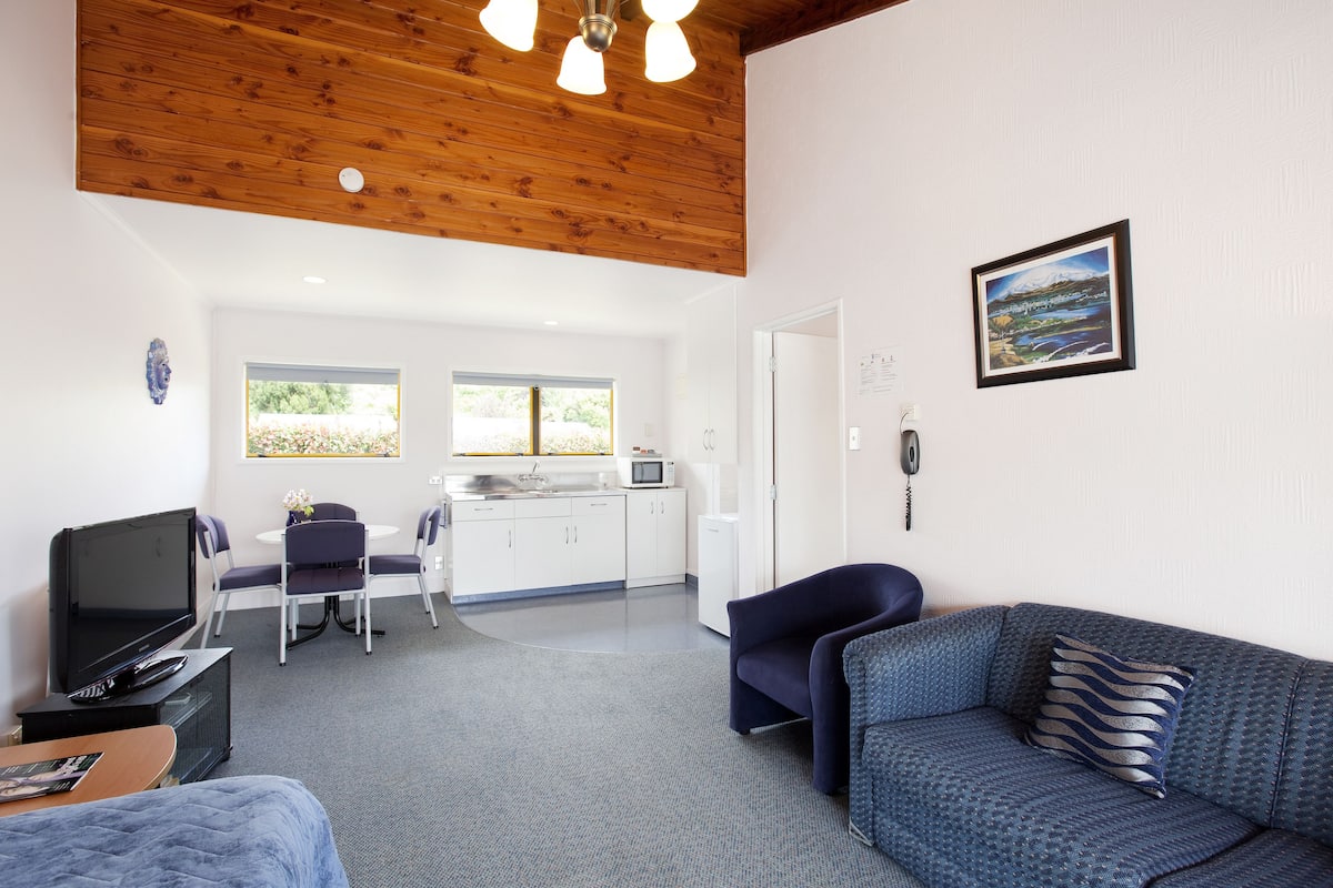 A modern hotel room with a wooden ceiling, a dining area with a table and chairs, a sofa, a TV, and a kitchen area.