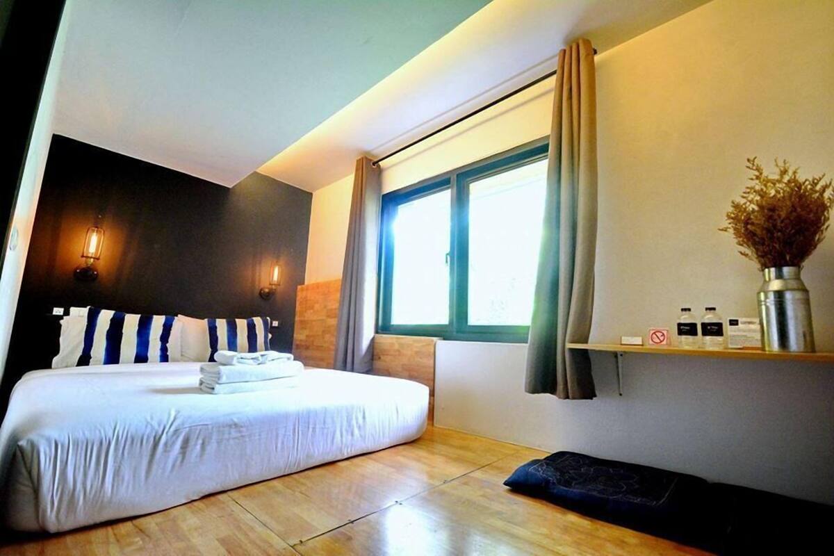 A hotel room with a large bed, a bedside table with toiletries, a window with curtains, and a wall-mounted shelf with items.