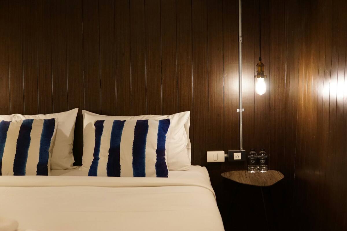 A hotel room with a wooden panel wall, a bed with white bedding and striped pillows, a bedside table with two water bottles, and a hanging light fixture.