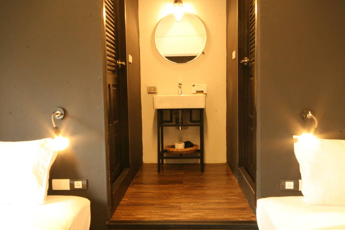 A hotel bathroom with a wooden floor, a round mirror, and two white beds with lights on.