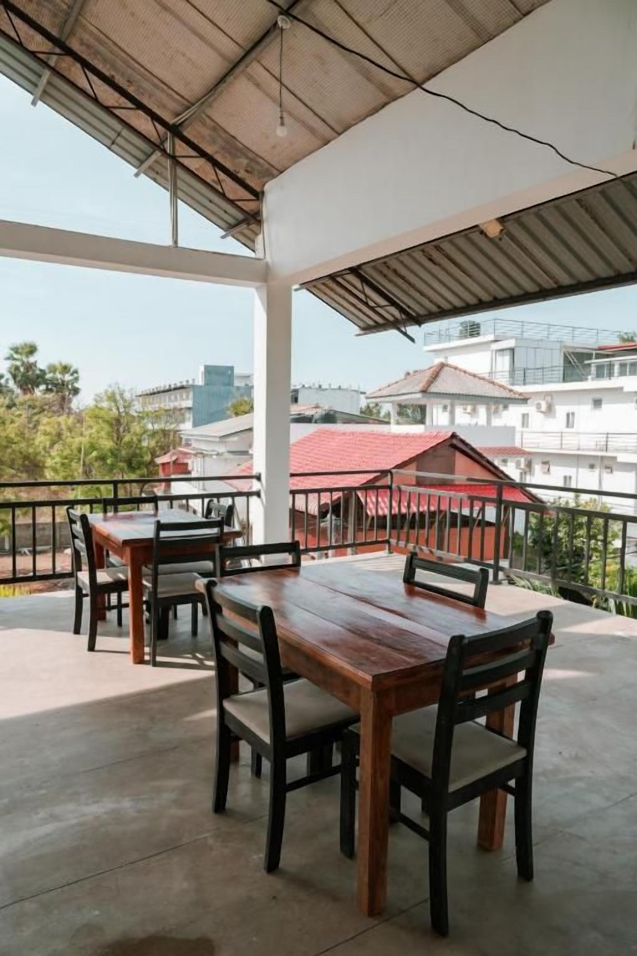 An outdoor patio with wooden tables and chairs, a metal railing, and a view of buildings in the background.