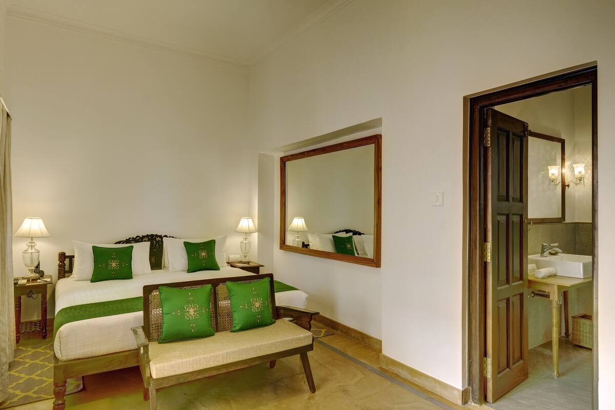 A hotel room with a bed, bedside tables, a bench, a large mirror, and a bathroom with a sink and mirror.