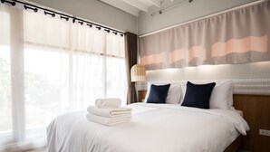 A neatly made bed with white linens and a folded towel, a bedside lamp, and a window with sheer curtains. - Thapae Happy House (Chiang Mai)