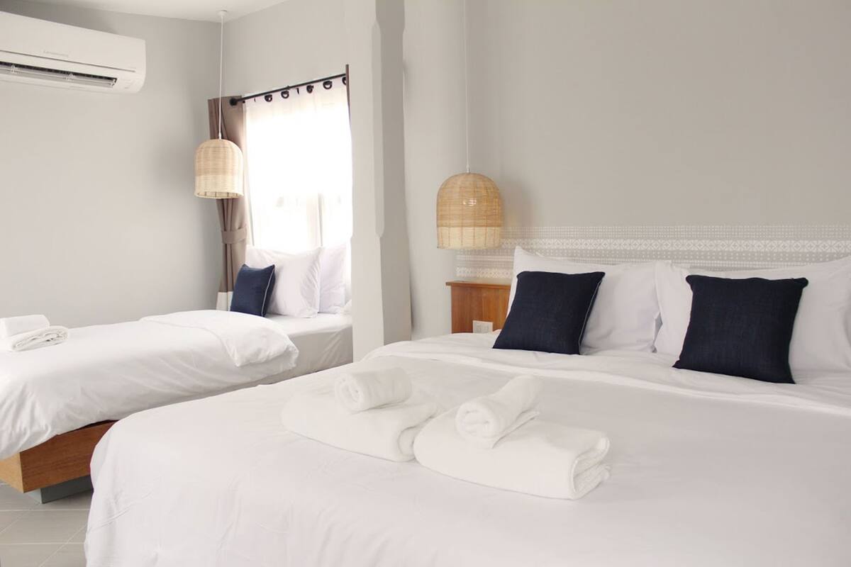A hotel room with two beds, white linens, a bedside table, a lamp, and an air conditioning unit.