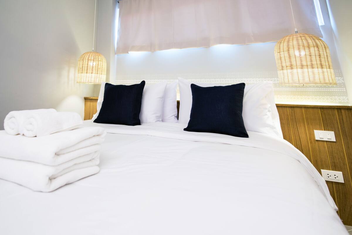 A neatly made bed with white linens and dark pillows, a stack of folded towels, and two wicker lamps.