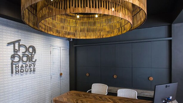 A modern office space with a herringbone wood table, a large hanging wooden light fixture, and a wall with the text "The House Happy House". - Thapae Happy House (Chiang Mai)
