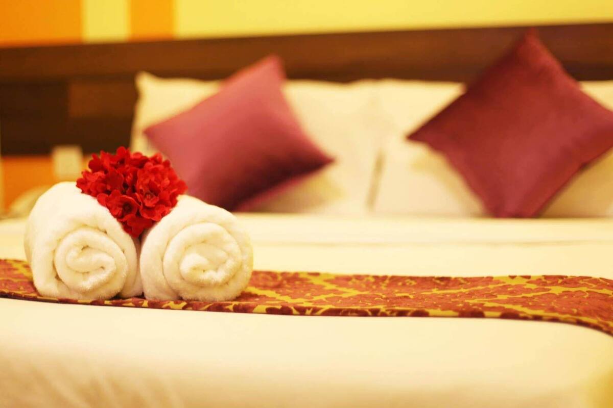 Una cama bien hecha con dos toallas blancas enrolladas y un arreglo floral rojo, sobre un edredón estampado y dos almohadas moradas.