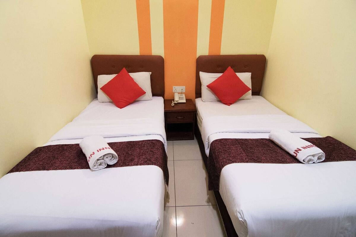 Dos camas individuales con almohadas rojas, una mesita de noche con un teléfono y una toalla con el logotipo de 'UN Inn'.