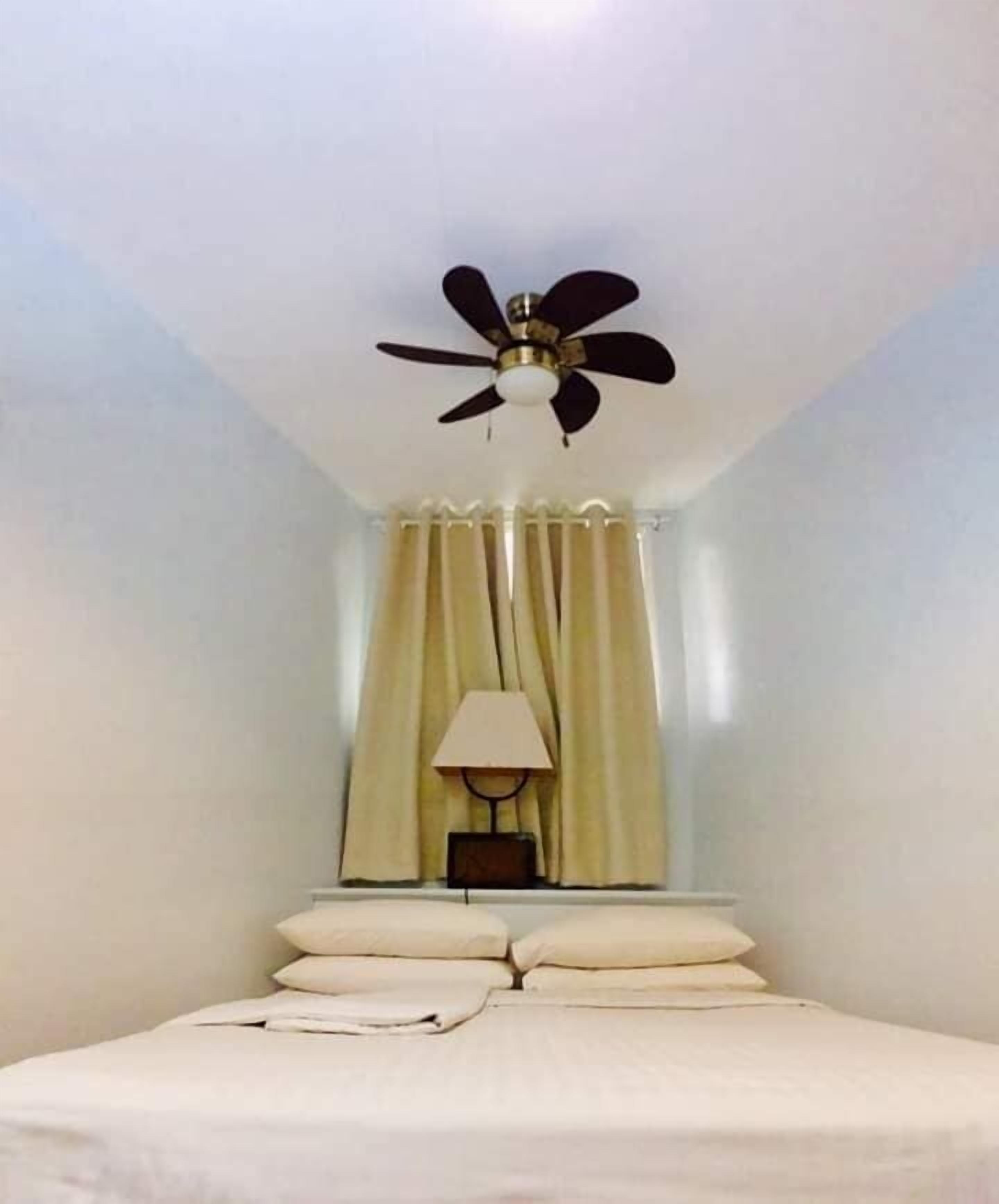 Una habitación de hotel con una cama, un ventilador de techo, una lámpara y cortinas.