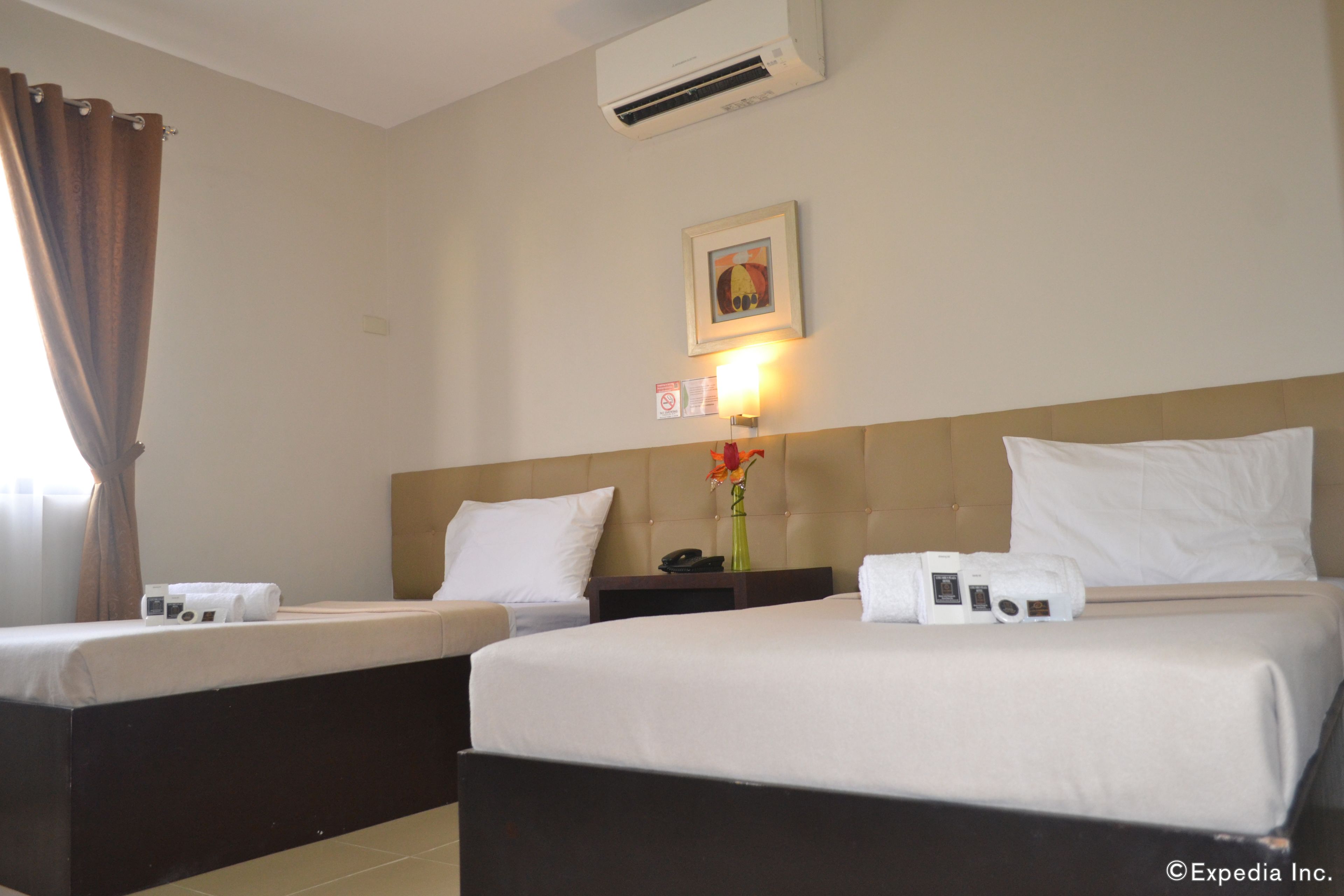 A hotel room with two beds, a nightstand, a wall-mounted air conditioner, a framed picture, and a wall-mounted light fixture.