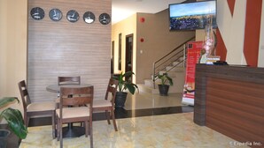 A hotel lobby with a reception desk, a TV mounted on the wall, a clock displaying time in four cities, and a staircase leading to another floor.
