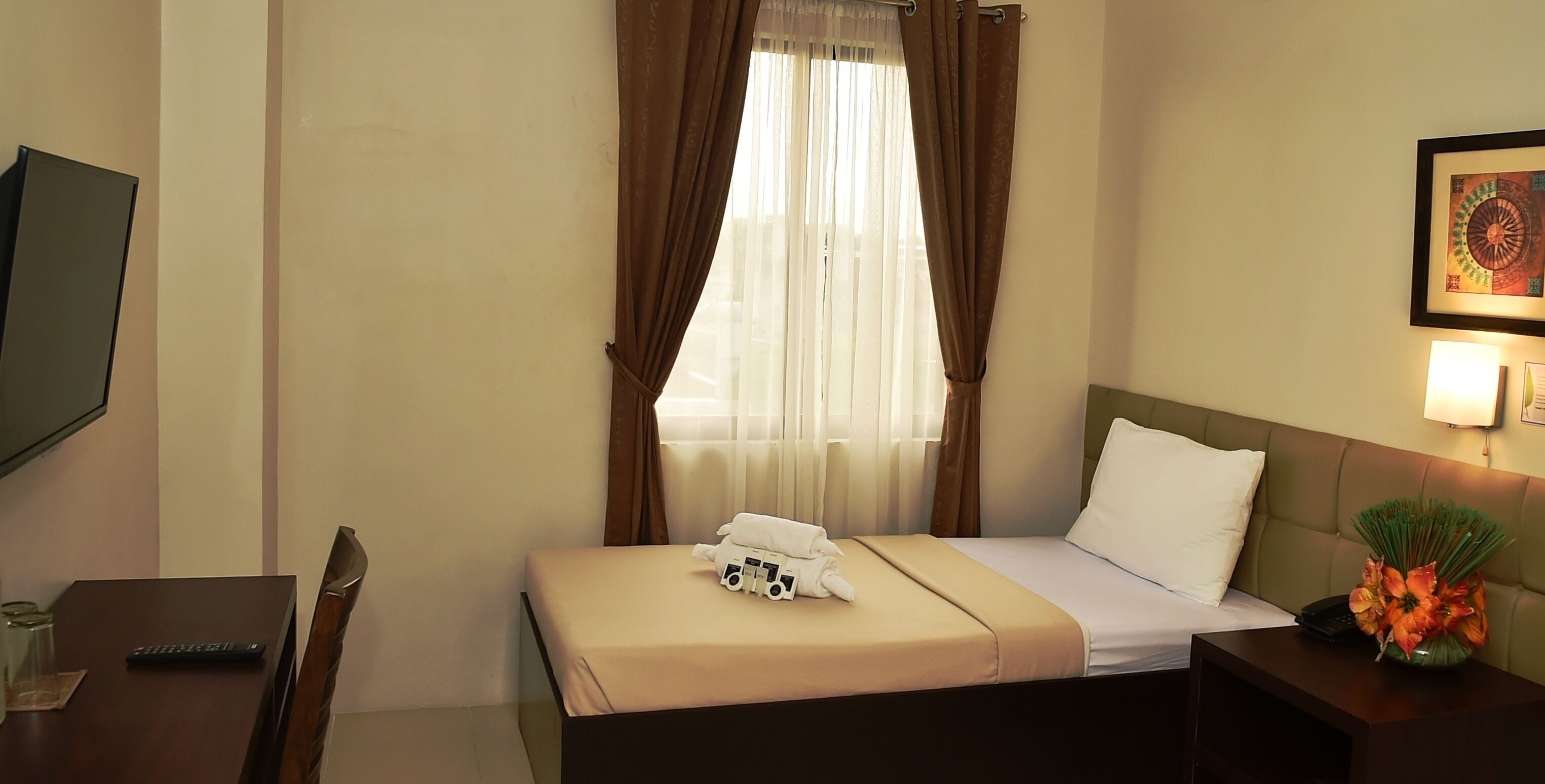 A hotel room with a bed, a bedside table with a lamp, a TV on the wall, and a window with curtains.