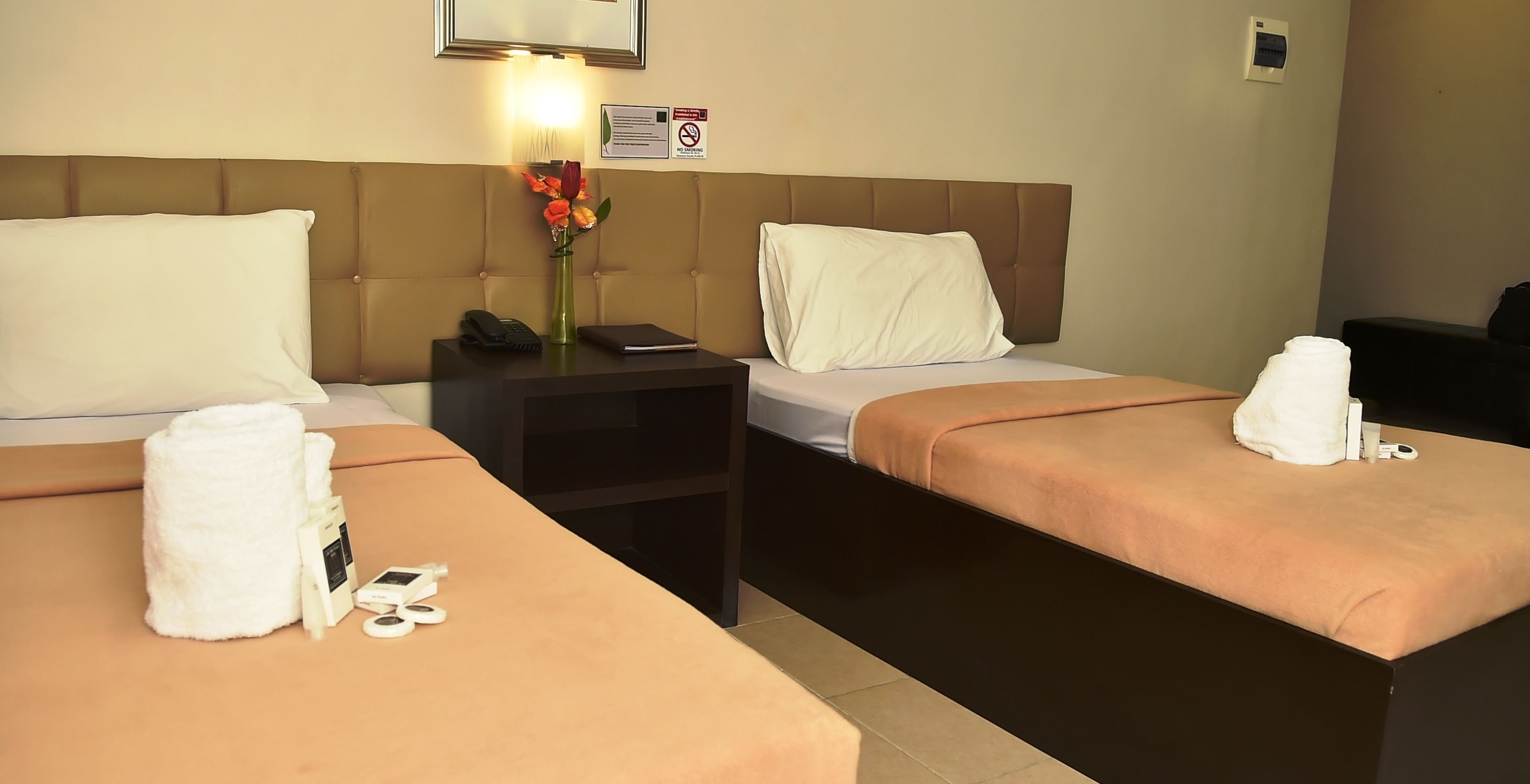 Habitación de hotel con dos camas, cada una con un almohadón blanco y cabecera marrón. Se observa un mesita de noche con un teléfono, un florero con flores y un cartel en la pared.
