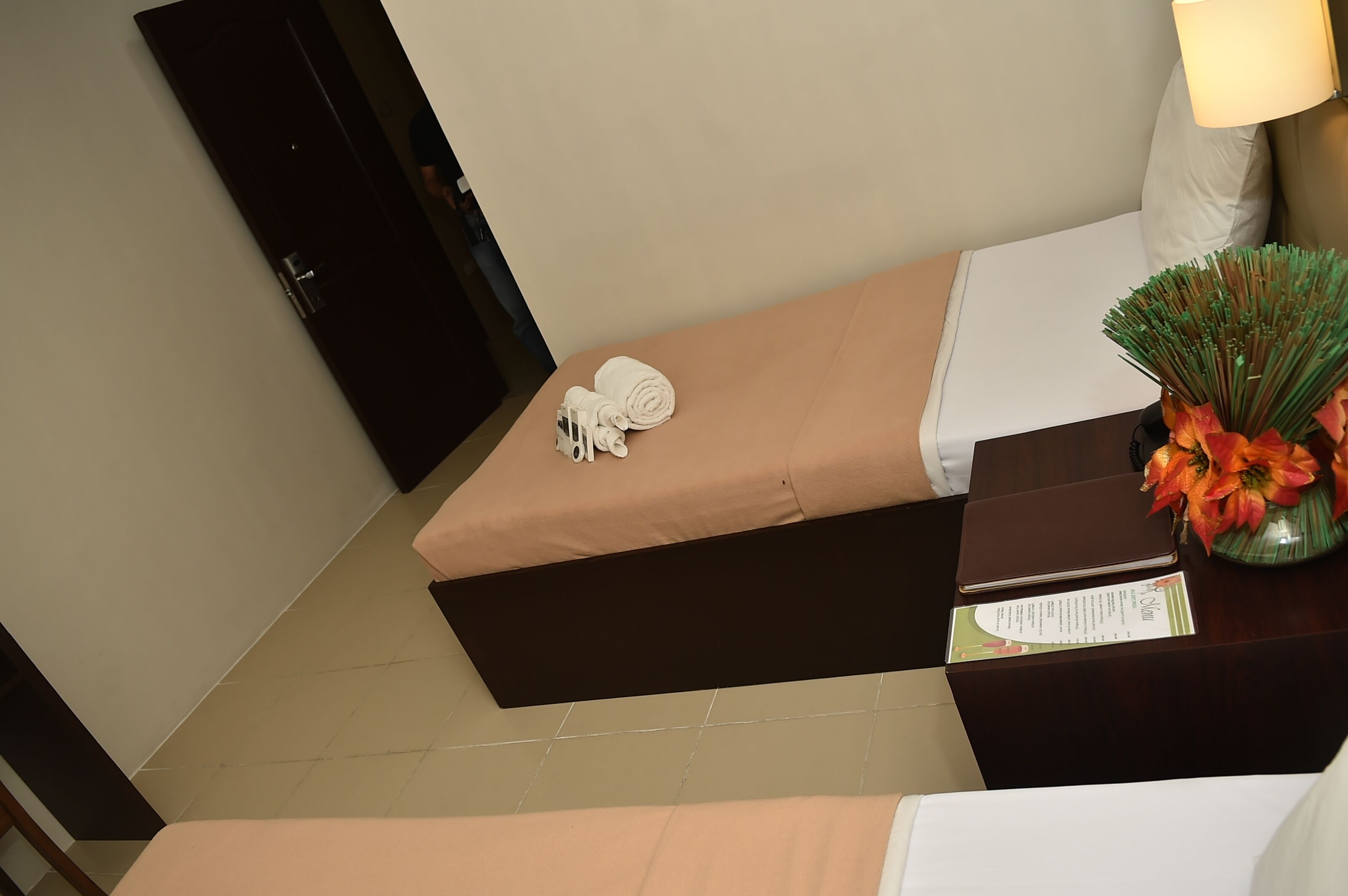 Una habitación de hotel con una cama, una mesita de noche con una lámpara, un jarrón con flores y un cartel sobre la mesita de noche.