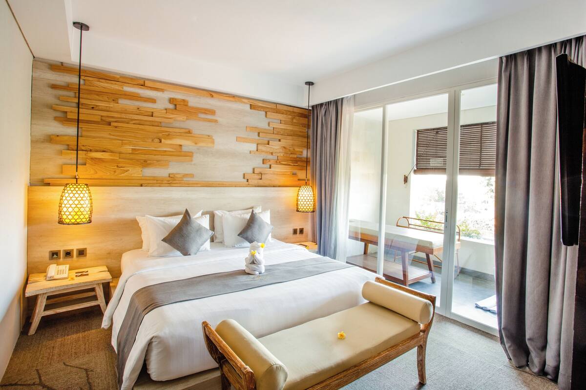 A modern hotel room with a large bed, a sofa, a wooden headboard, and a balcony with a table and chairs.