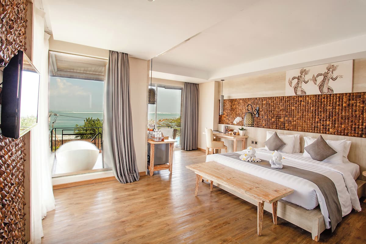 A spacious bedroom with a large bed, a wooden coffee table, and a desk with a chair. There is a balcony with a view of the sea.