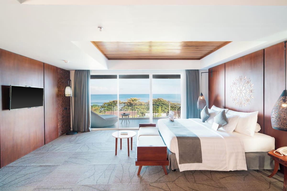 A modern hotel room with a large bed, a TV, a balcony with a view of the ocean, and a small seating area.