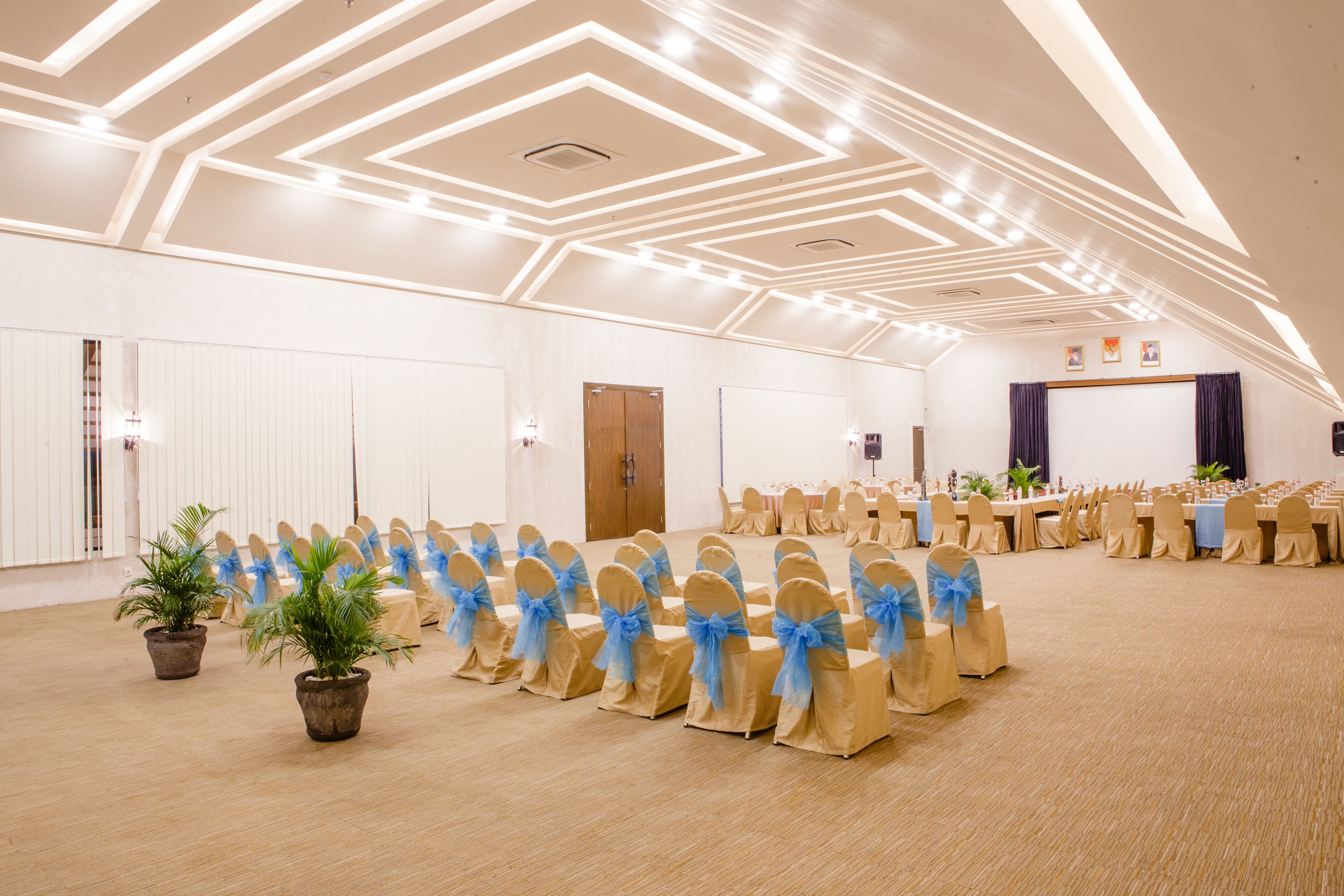 A large hall with rows of chairs and tables, a stage, and a high ceiling with geometric lighting.