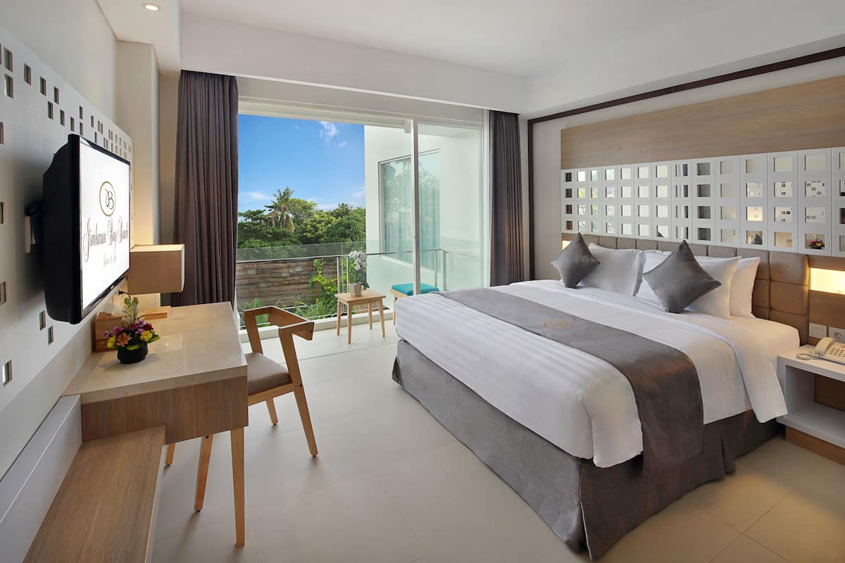 A modern hotel room with a large bed, a desk with a chair, a TV, and a balcony with a view of the outdoors.