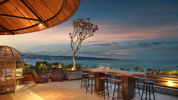 A rooftop bar with wicker chairs and stools, overlooking a scenic view of the sea and mountains. - Jimbaran Bay Beach Resort & Spa (Kedonganan)