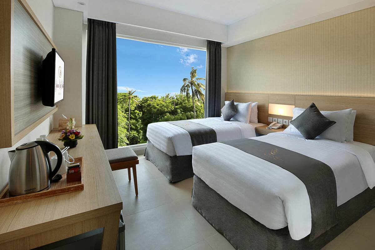 A hotel room with two beds, a desk with a kettle, a telephone, and a window with a view of palm trees.