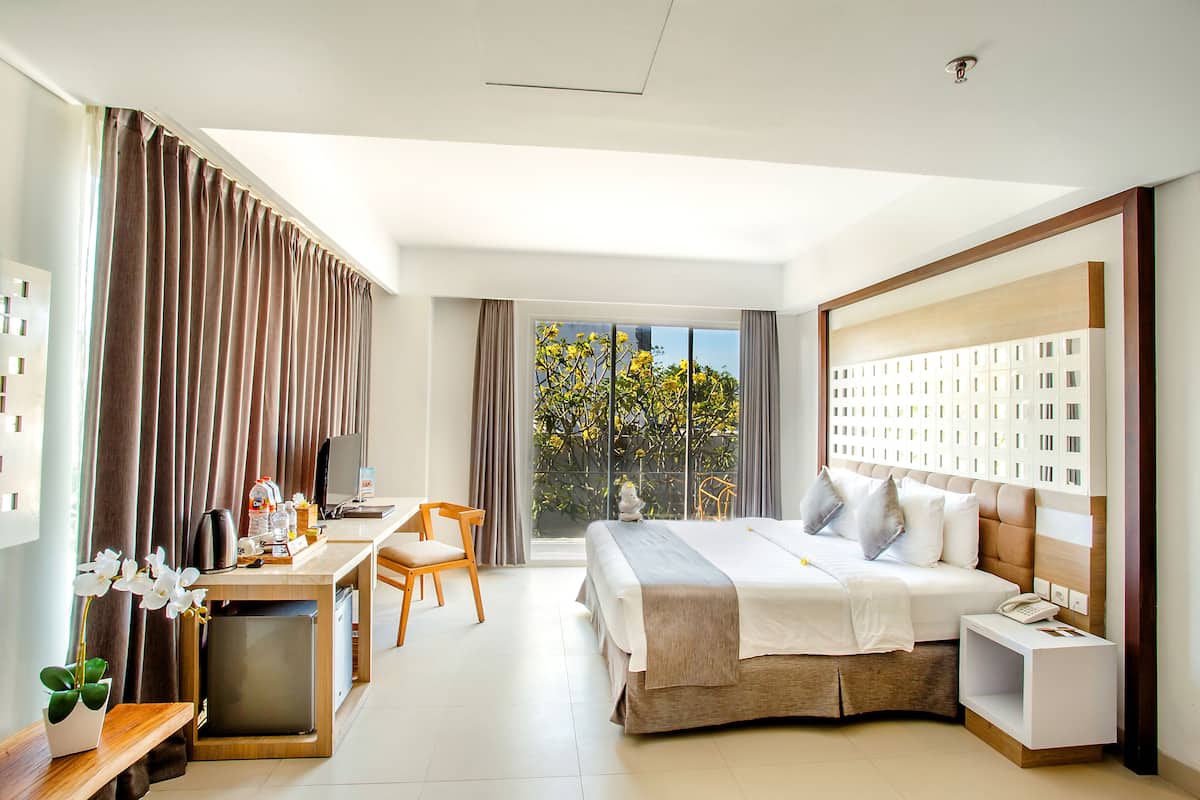 A hotel room with a bed, desk, chair, and a view of greenery outside.