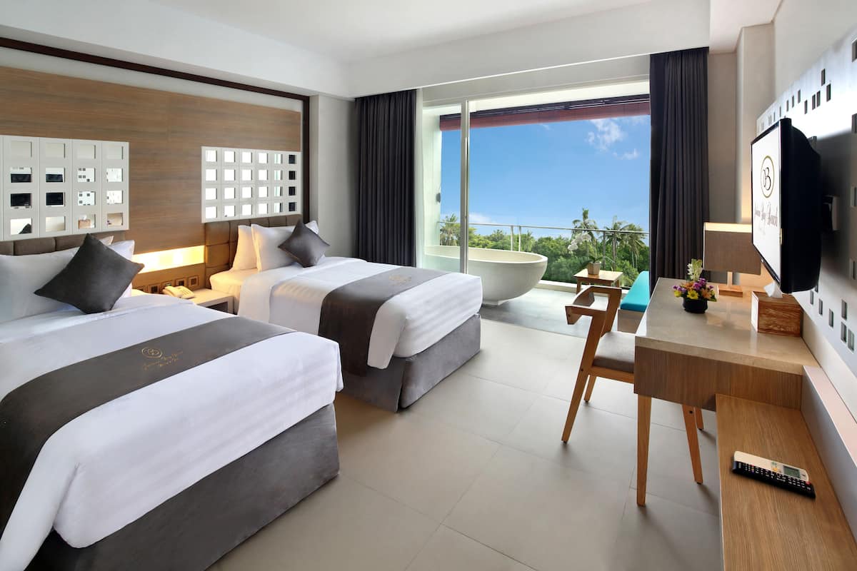 A hotel room with two beds, a desk, a TV, and a balcony with a view of the ocean.