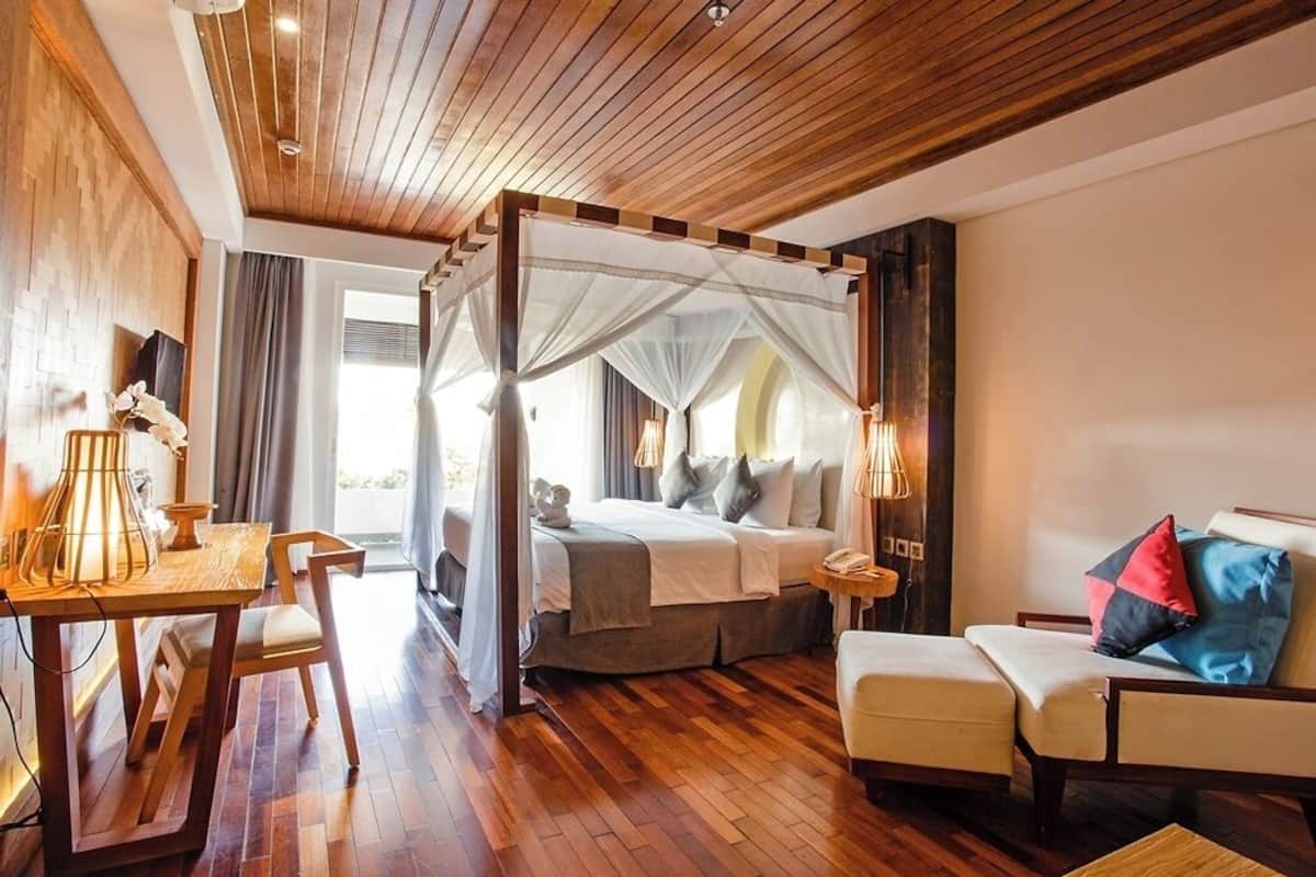 A bedroom with a four-poster bed, a sitting area with a sofa and armchair, a small table with a lamp, and a view of the outdoors.