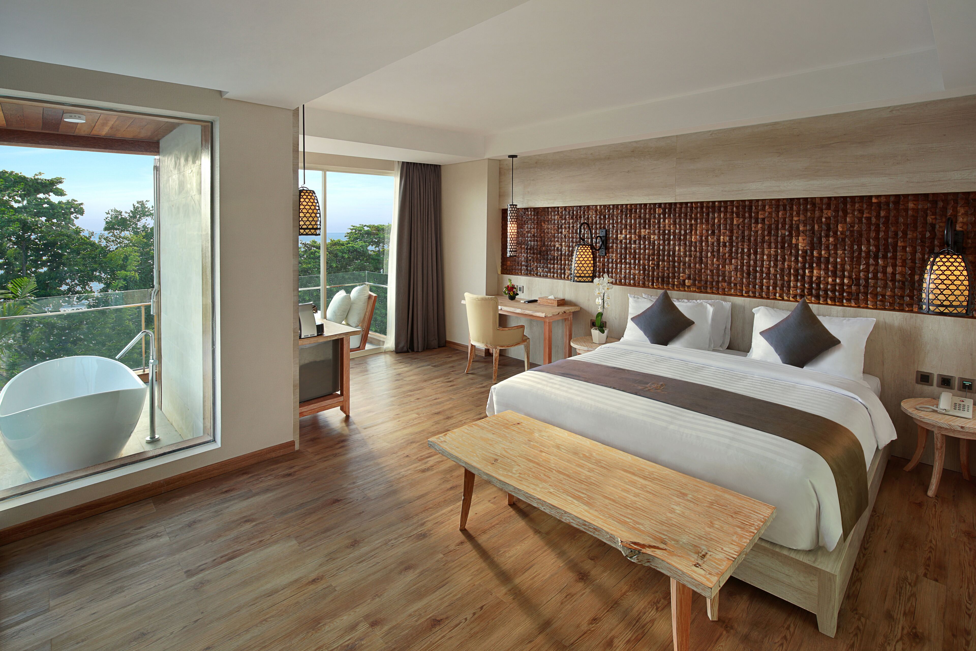 Een moderne hotelkamer met een groot bed, een houten bankje, een bureau en een balkon met uitzicht op de bomen.