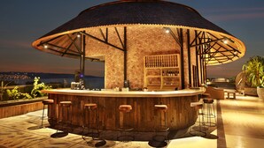 An outdoor bar with a wooden structure, bar stools, and a view of the city at dusk. - Jimbaran Bay Beach Resort & Spa (Kedonganan)