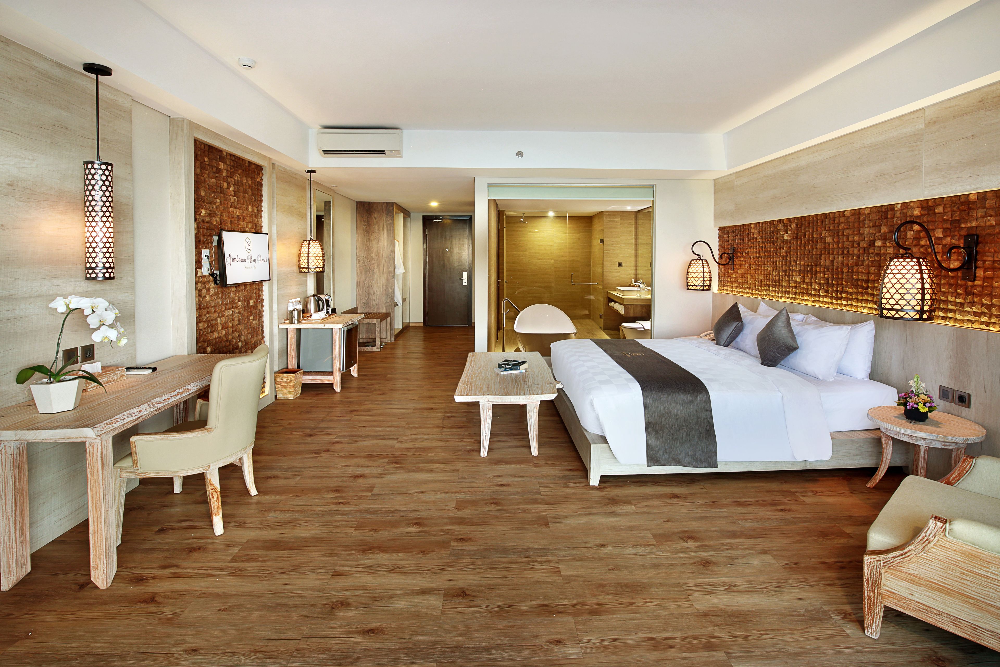 A modern hotel room with a wooden floor, a bed with white bedding, a desk with a chair, a small table, and a bathroom visible through an open door.