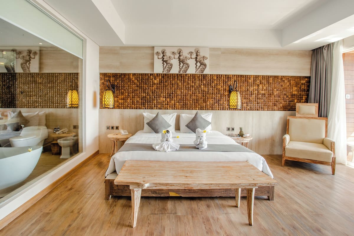 A modern hotel room with a large bed, a wooden bench, a wicker chair, and a view of the bathroom.