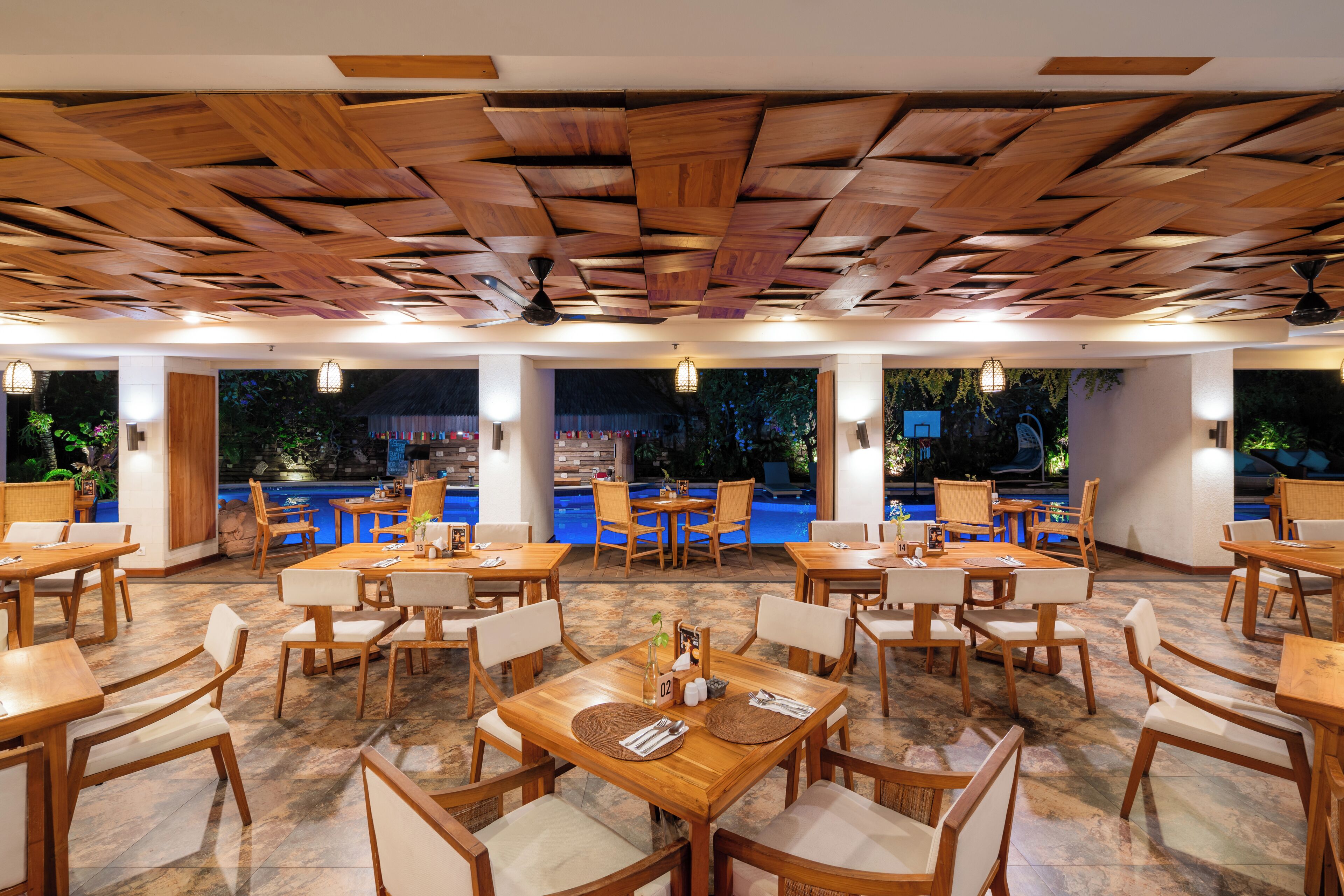 Restaurante com mesas e cadeiras de madeira, piscina ao fundo e teto com padrão geométrico.
