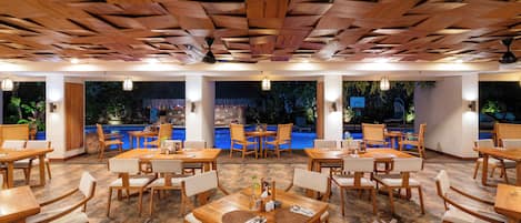 Restaurante com mesas e cadeiras de madeira, piscina ao fundo e teto com padrão geométrico.