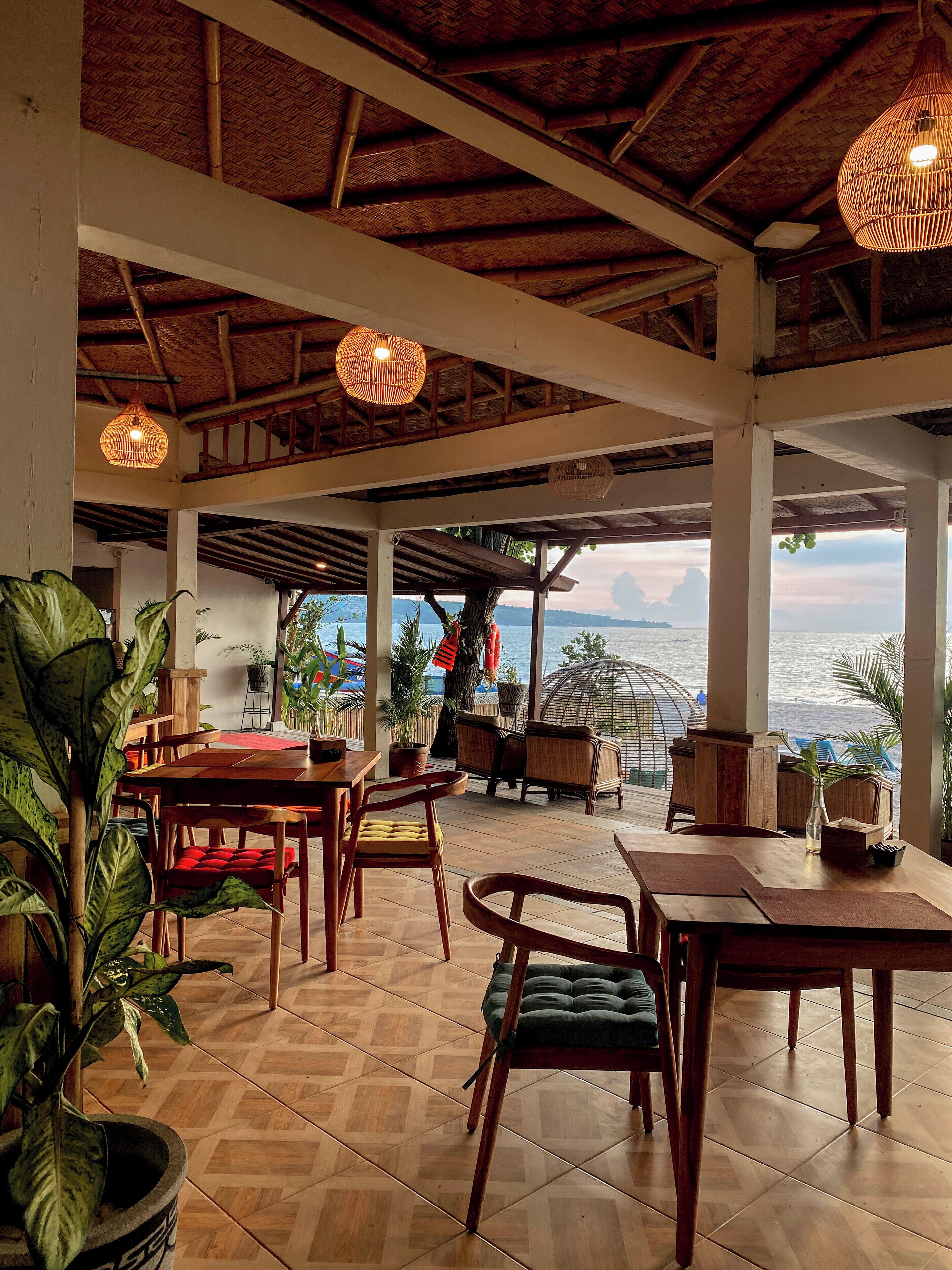 Un comedor interior con mesas y sillas de madera, plantas en macetas y vistas al mar.