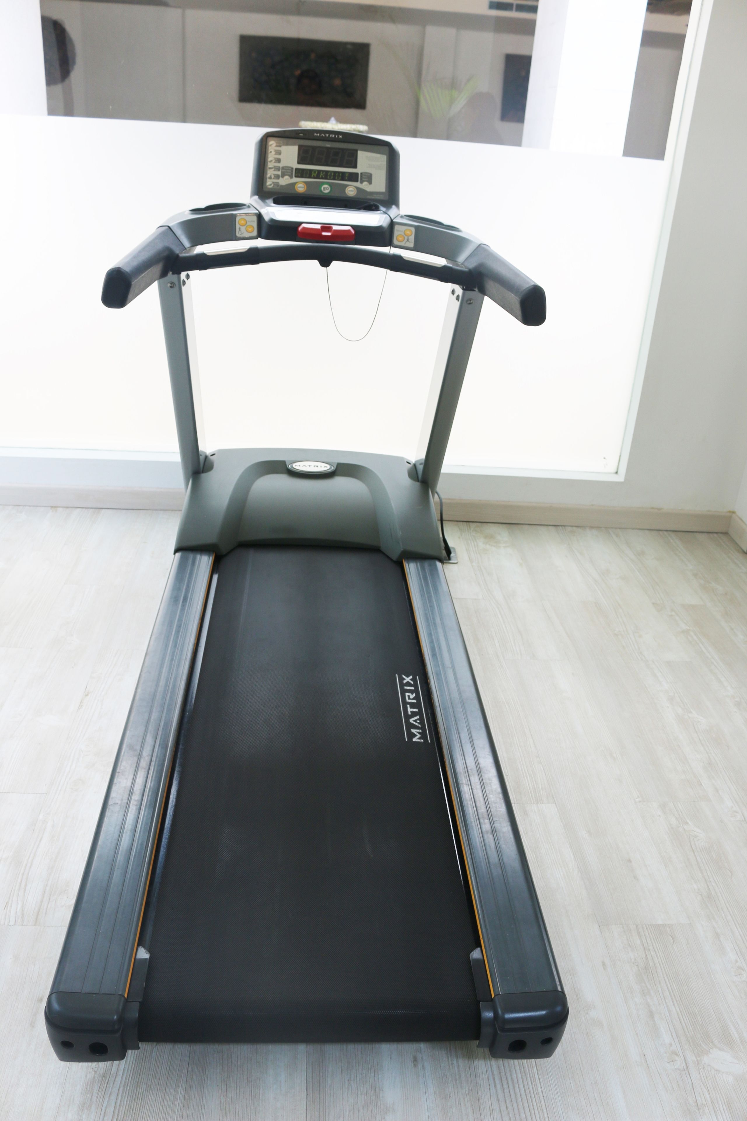 Un tapis de course dans une pièce dotée d’une grande fenêtre, d’un plancher en bois clair et d’une porte bien visible.