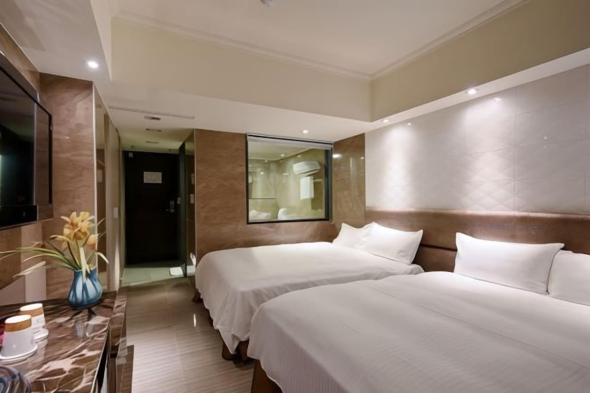 Una habitación de hotel con dos camas, un televisor, un jarrón con flores y un espejo.