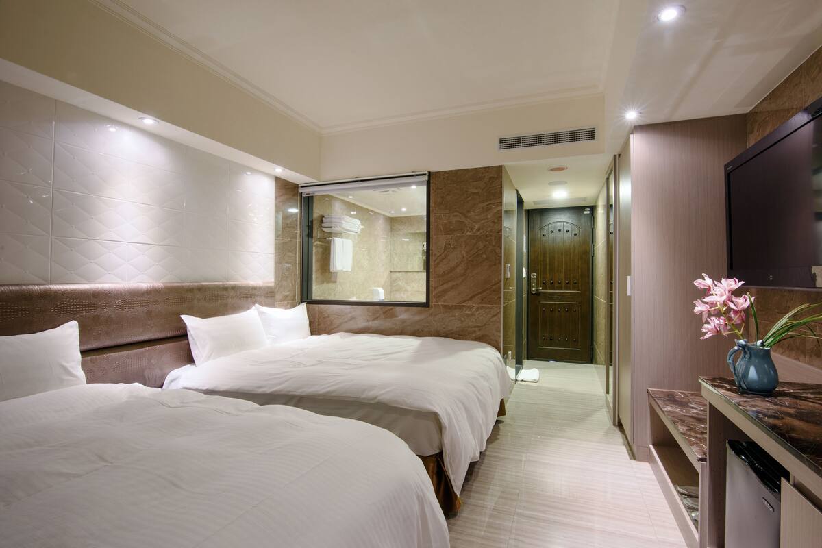 A hotel room with two beds, a bathroom with a glass shower, and a small table with a vase of flowers.